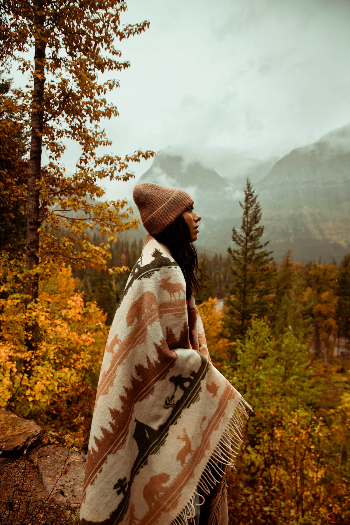 Person wrapped in a patterned blanket standing in a forest with mountains in the background