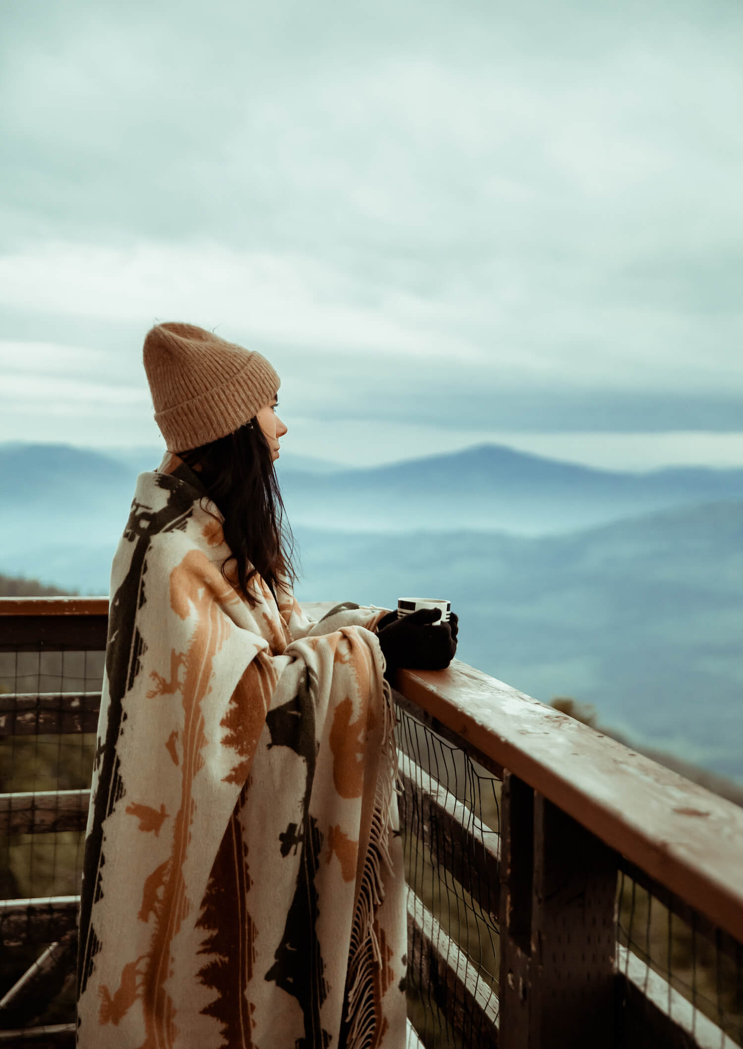 Person wrapped in a patterned blanket, wearing a beanie, looking out over a mountain range.