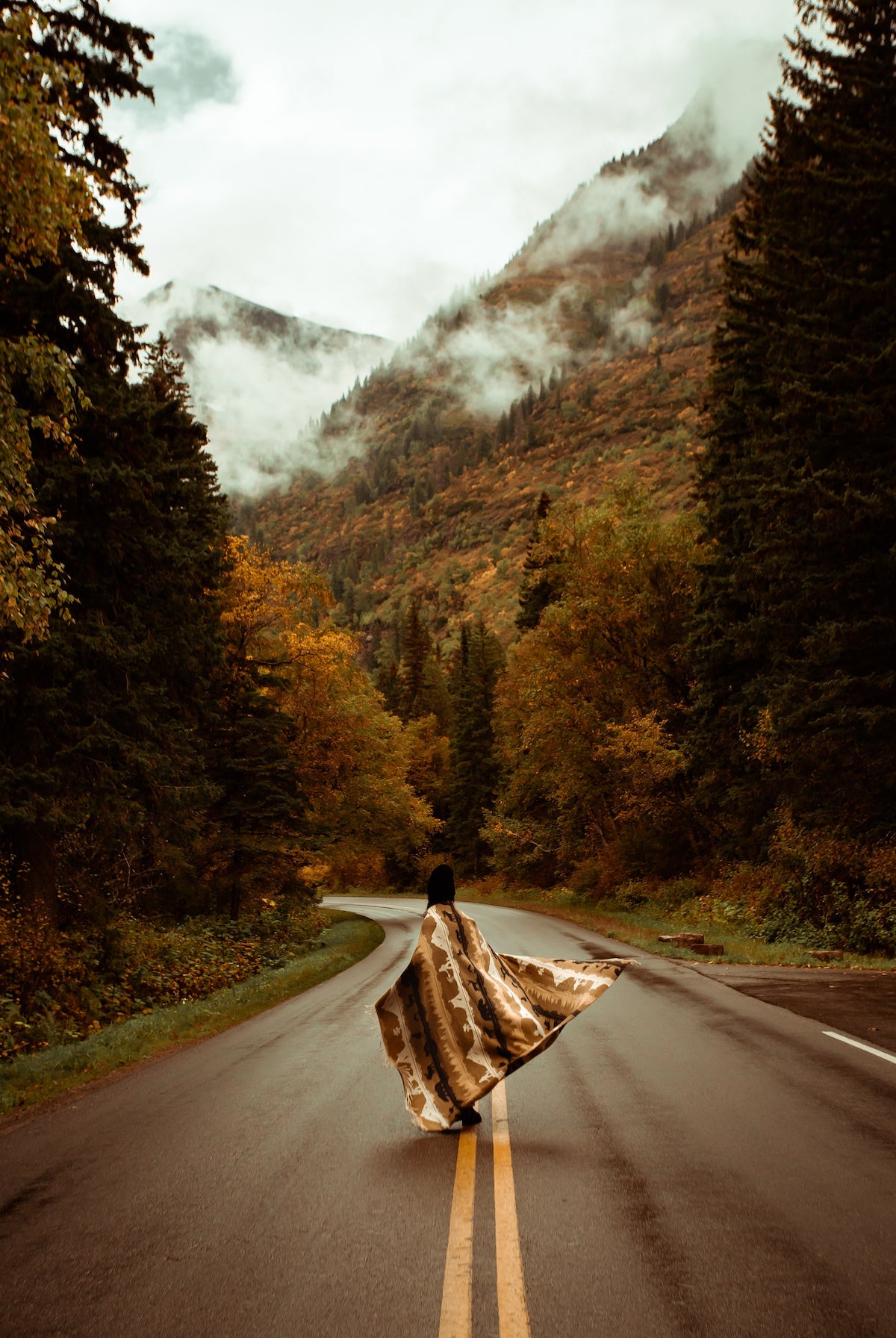 Person with a patterned blanket standing in the middle of a scenic mountain road.