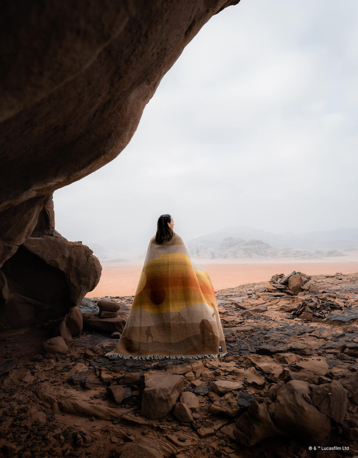 Person wrapped in a colorful blanket standing inside a cave with a desert landscape outside