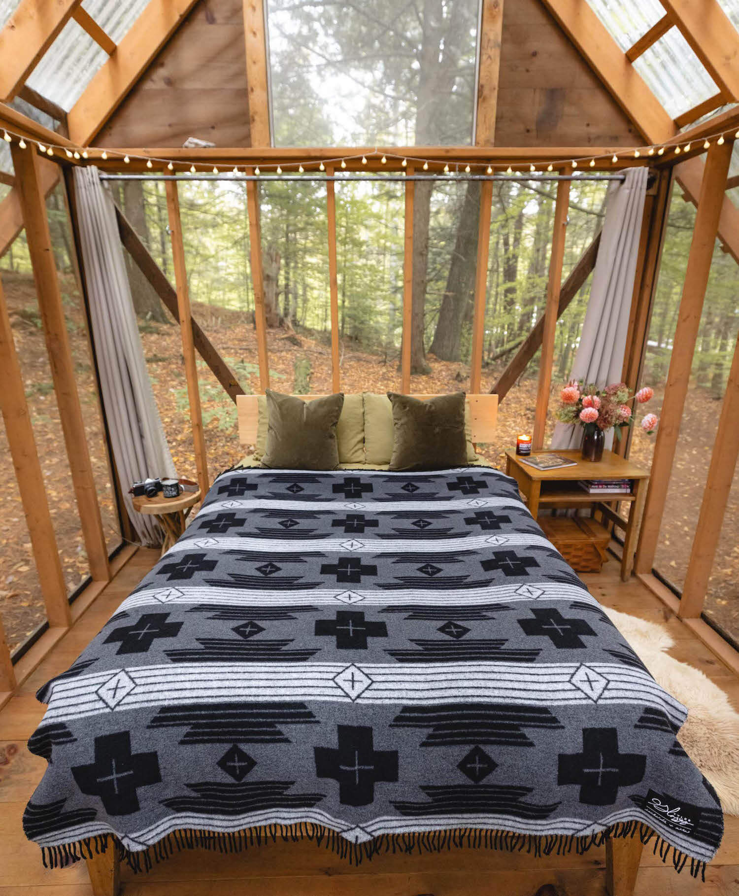 Bed with patterned blanket in a wooden cabin with large windows overlooking a forest.