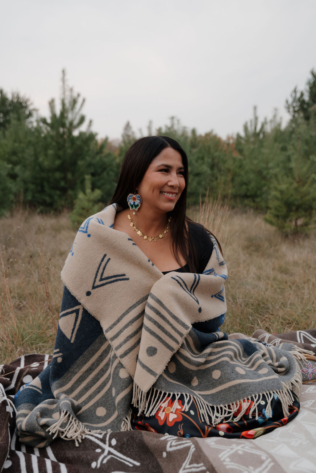 Woman sitting outdoors with a patterned blanket draped over her, surrounded by trees.