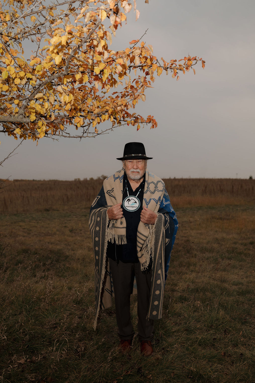 Man in a hat and coat holding a patterned scarf in a field with a tree in the background